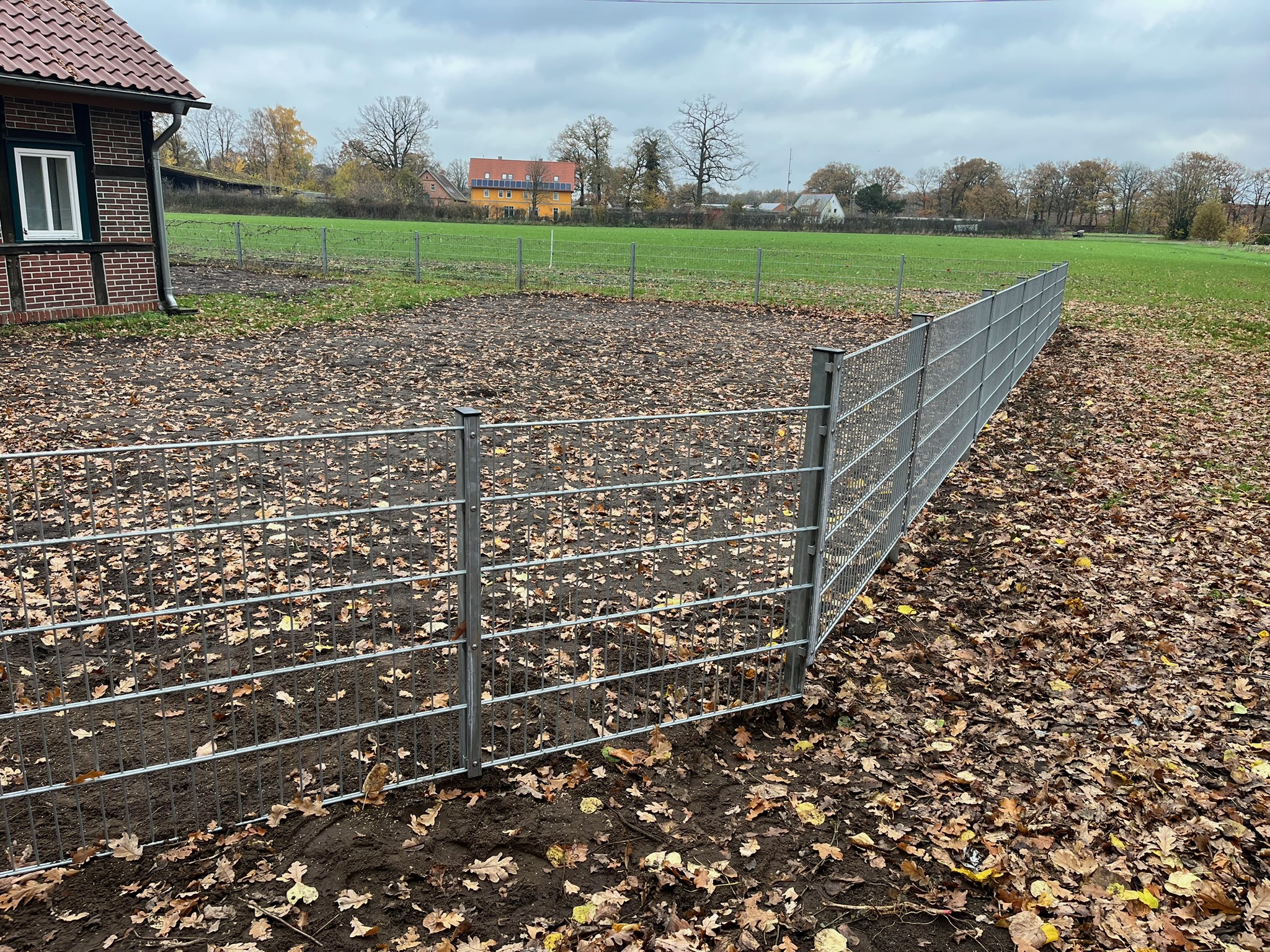 Ein frisch aufgestellter, verzinkter Doppelstabmattenzaun grenzt einen Gartenbereich mit laubbedecktem Erdboden professionell ein. Links ist ein Teil eines traditionellen Backsteingebäudes zu sehen, während sich im Hintergrund weite grüne Wiesen unter einem herbstlich bewölkten Himmel erstrecken.