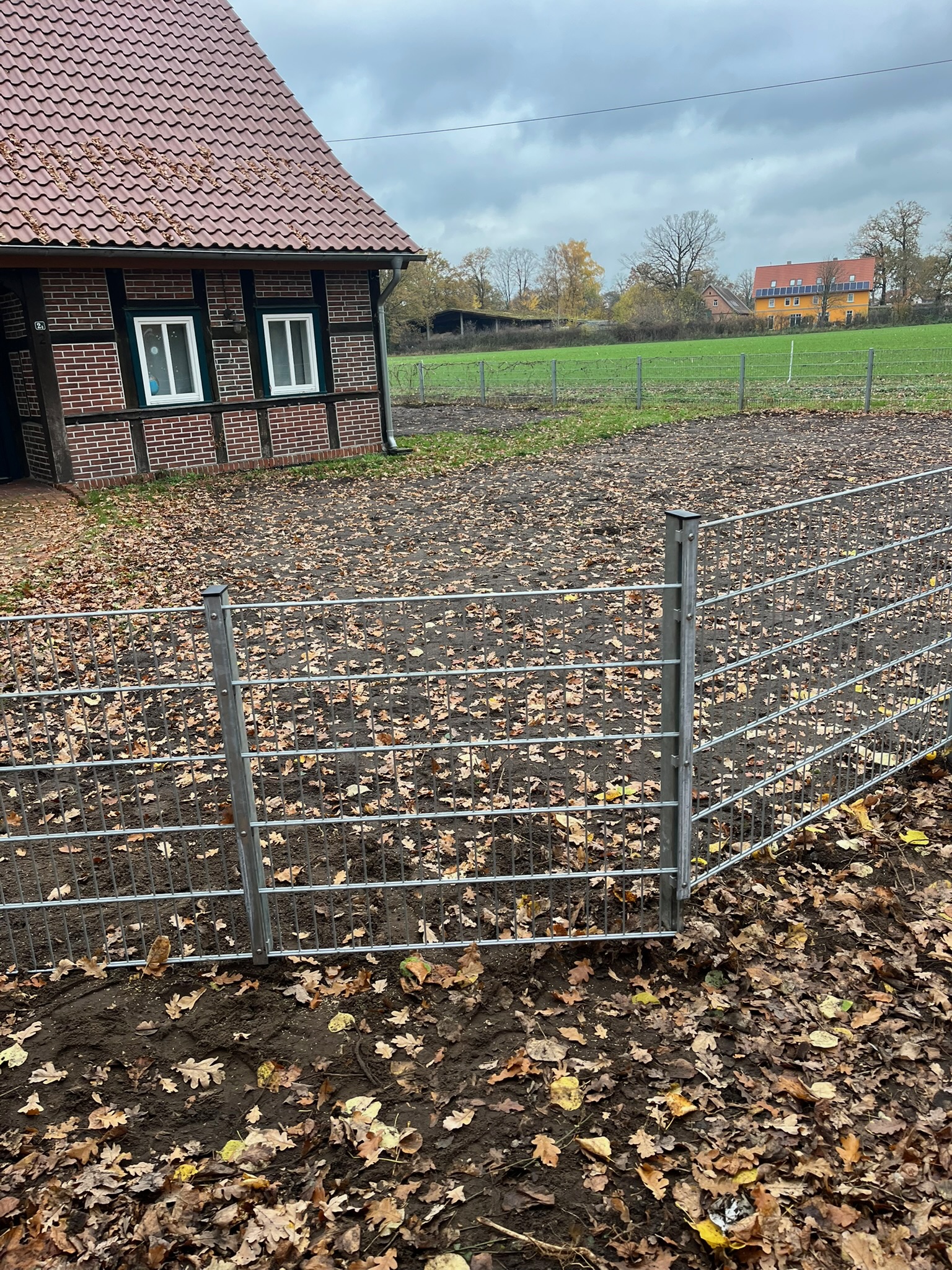 Ein fachgerecht montierter, verzinkter Doppelstabmattenzaun grenzt einen laubbedeckten Gartenbereich ab. Im Hintergrund links ist ein klassisches Fachwerkhaus aus rotem Backstein zu sehen, während sich rechts eine weite, grüne Wiese unter einem bewölkten Herbsthimmel erstreckt.